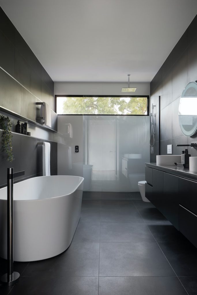 Modern bathroom with freestanding tub, dark tiles, large mirror, and natural light through a wide window