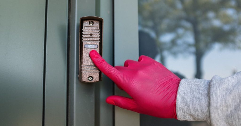 A red-gloved hand presses a modern doorbell on a metal door, depicting security and service